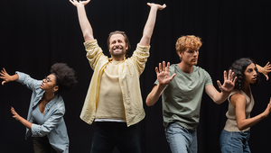 Vier junge Erwachsene stehen vor schwarzem Hintergrund in expressiven Theaterposen – mit erhobenen Armen, ausdrucksstarken Gesichtern und offenen Gesten. Das Bild zeigt vier junge Erwachsene, die in einer Reihe nebeneinander vor einem schwarzen Bühnenhintergrund stehen. Sie befinden sich in verschiedenen, theatralisch überzeichneten Posen, als wären sie mitten in einer Improvisations- oder Schauspielübung. Ganz links beugt sich eine Frau mit dunklem Lockenhaar und Brille leicht nach vorne, die Arme dramatisch ausgestreckt. Daneben steht ein Mann mit mittellangen Haaren und Bart, der strahlend lächelt und beide Arme siegreich nach oben reißt. Der dritte Darsteller, ein junger Mann mit rotem Lockenkopf, hebt die Hände in einer abwehrenden Geste, sein Gesicht wirkt ernst und konzentriert. Ganz rechts schaut eine junge Frau mit dunklem, gewelltem Haar aufmerksam nach vorn, die Arme leicht angewinkelt, als würde sie sich vorsichtig vortasten. Alle tragen legere Kleidung und wirken fokussiert und engagiert. Die Szene vermittelt Energie, Ausdruckskraft und Spielfreude – typisch für Theater- oder Schauspieltrainings.